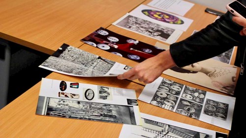 Image shows a curatorial activity with printed images of artworks on a table being arranged by hands coming in from the right