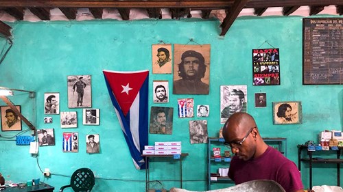 Image shows a young Cuban man with glasses sitting with what look to be large weighing scales. He is in front of a turquoise wall with a Cuban flag and images of Che Guevara and a young Fidel Castro.