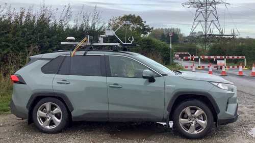 A grey four-wheel drive car with equipment mounted on the roof-rack parked in a layby near an electricity pylon
