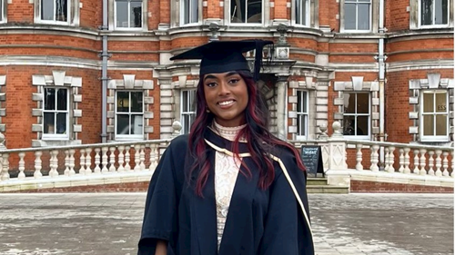 Thulasi, alumni, wearing graduation gown in front of Founders Building