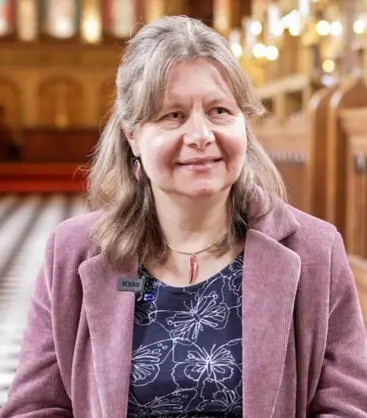 Head shot of Professor Hannah Thompson, pictured in the Royal Holloway chapel, wearing a soft-textured purple blazer over a blue top with large butterflies in white outline. She has long light-brown hair.