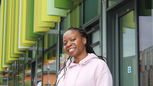 A headshot of PGT student Faith from Malawi smiling at camera. Faith has a dark skin tone and dark hair, is wearing a pink hoodie and is standing in front of the Moore building on campus