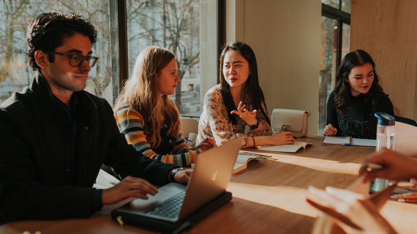 Four students seated around a wooden table, discussing with a laptop and papers.
